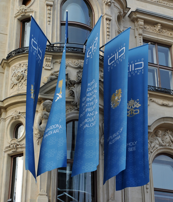 Bildunterschrift: Flaggen für die Unterstützer des Zentrums an der Fassade: (v.l.n.r.) Königreich Spanien, Königreich Saudi-Arabien, (das Zentrum selber), Republik Österreich, der „Heilige Stuhl“.