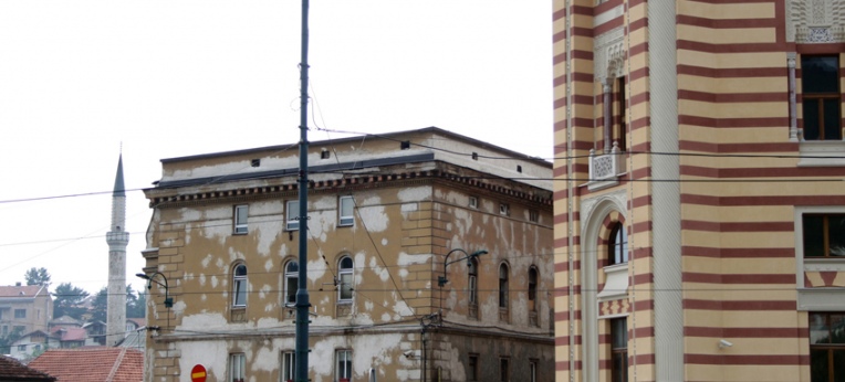 Neben der frisch renovierten Vijećnica verwittert ein Wohngebäude vor sich hin. Im Hintergrund das Minarett einer frisch renovierten Moschee.